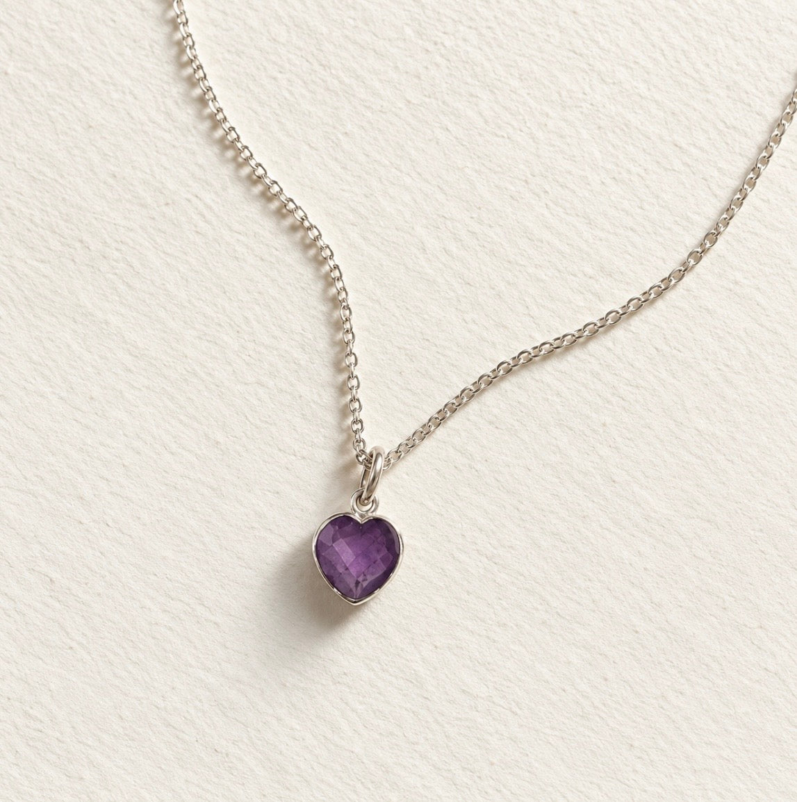 A silver chain necklace with a heart-shaped purple amethyst gemstone pendant, displayed on a textured off-white background.