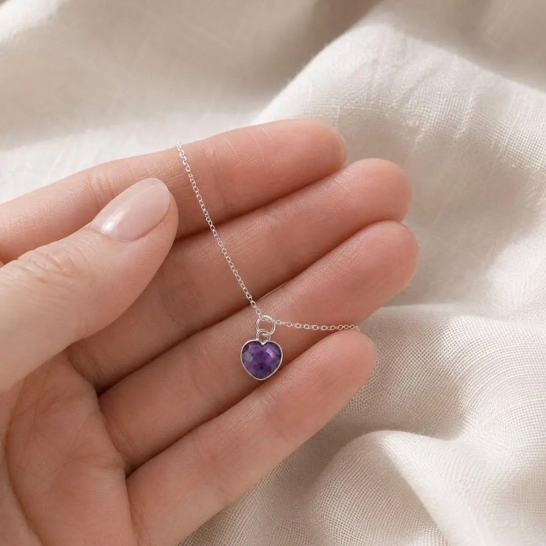A close-up of a hand holding a delicate silver chain necklace with a small heart-shaped purple amethyst gemstone pendant, set against a soft, draped white fabric background.