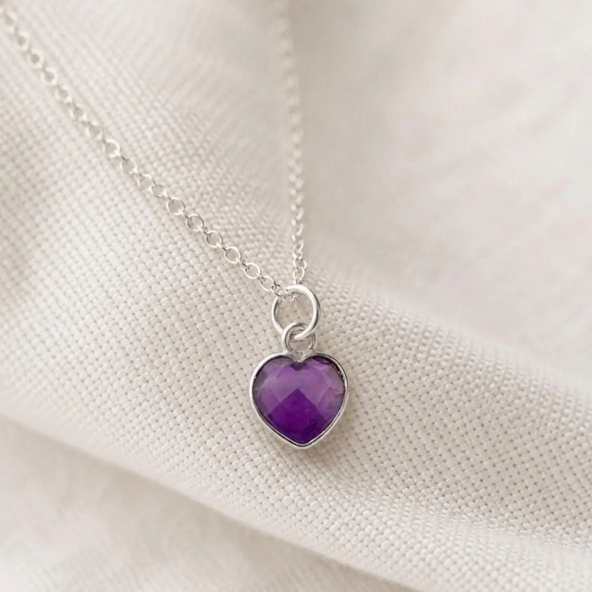 A silver heart-shaped pendant necklace with a faceted purple amethyst gemstone, resting on a textured off-white fabric background.