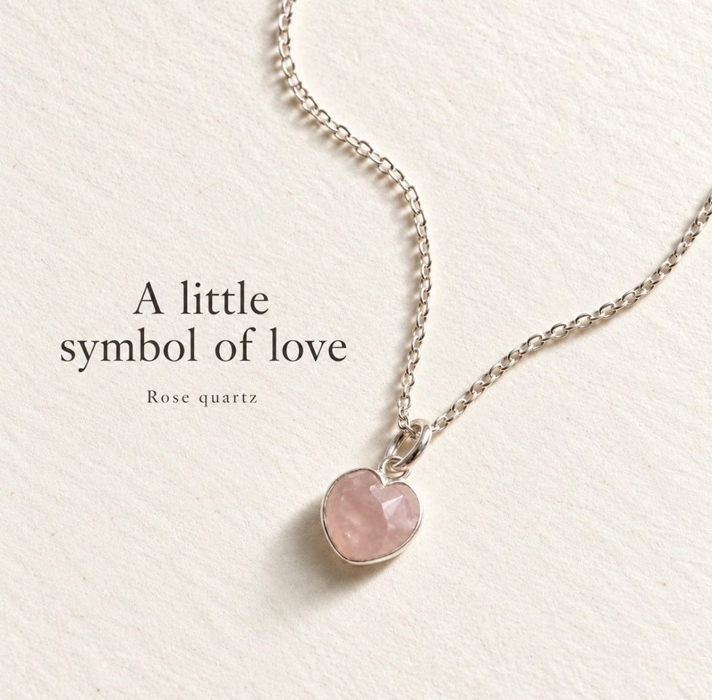 A heart-shaped rose quartz pendant on a delicate silver chain, displayed on a textured cream background with the text 'A little symbol of love Rose quartz'.