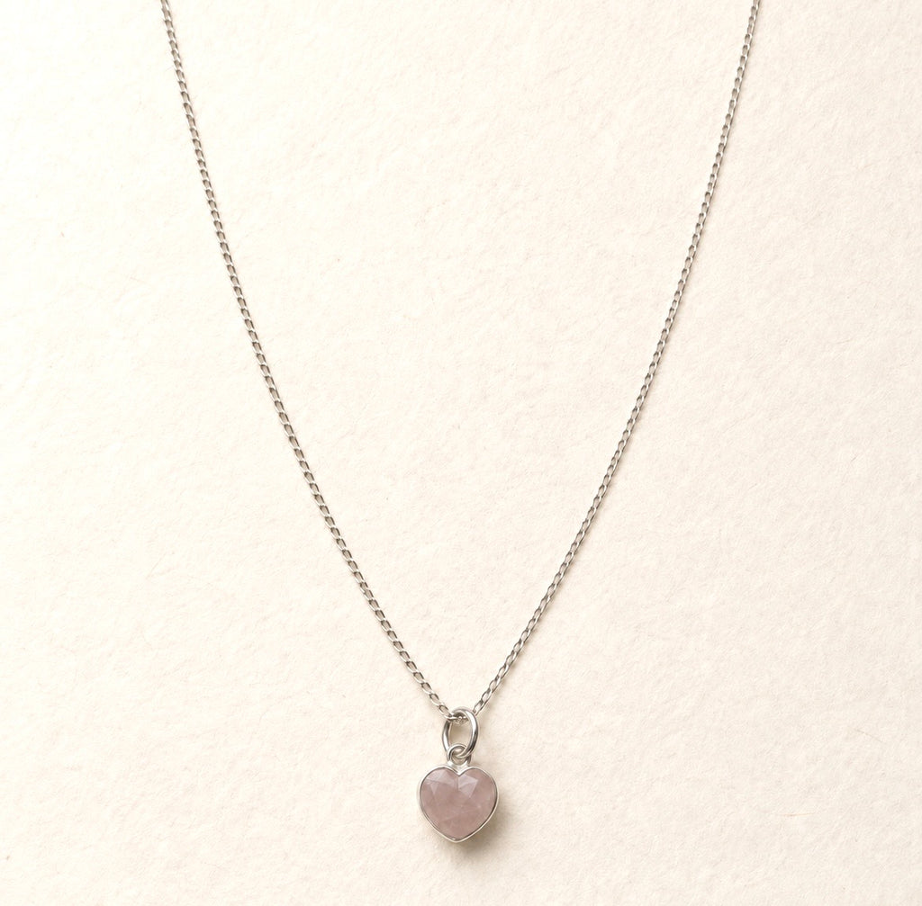 A delicate silver chain necklace with a small heart-shaped pink rose quartz stone pendant, displayed on a textured off-white background.