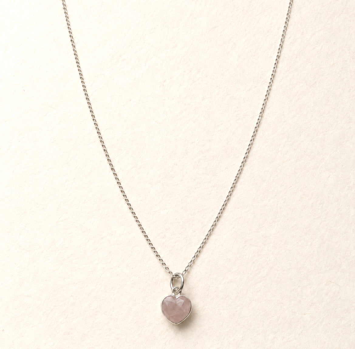 A delicate silver chain necklace with a small heart-shaped pink rose quartz stone pendant, displayed on a textured off-white background.
