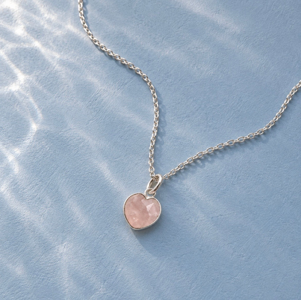 A delicate silver chain necklace with a small heart-shaped pink rose quartz stone pendant, displayed on a textured light blue surface with soft, dappled light reflections.