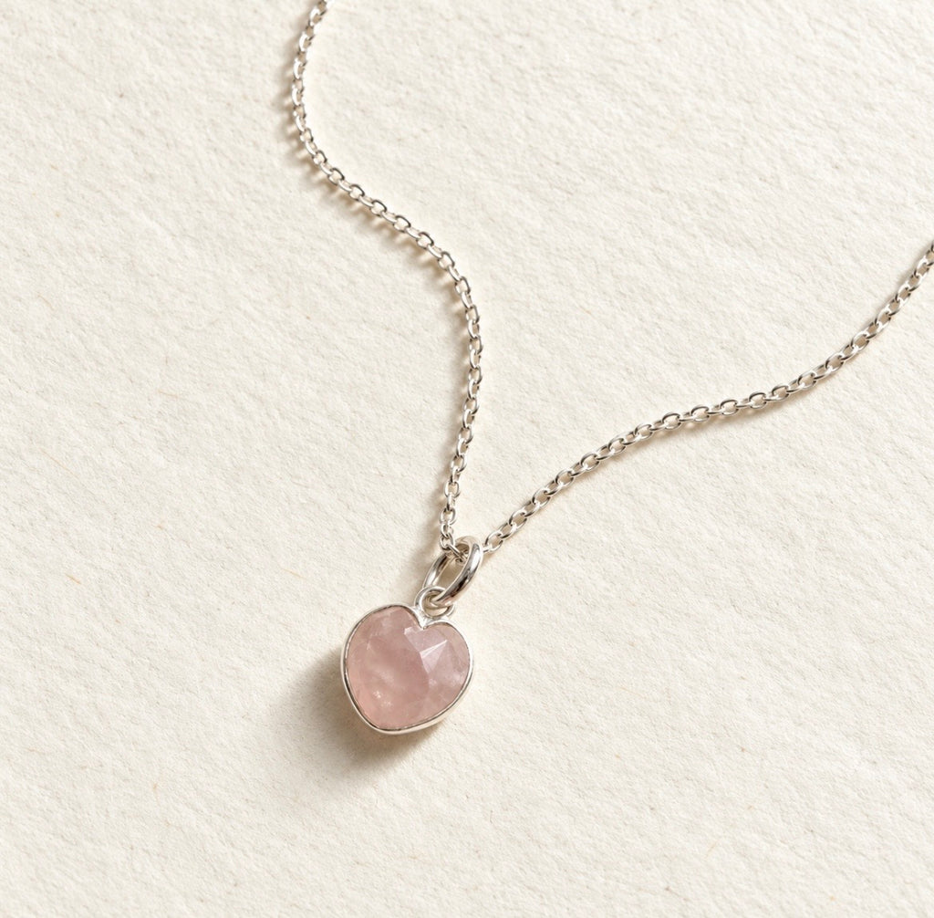 A delicate silver chain necklace with a small heart-shaped pink rose quartz gemstone pendant, displayed on a textured off-white background.
