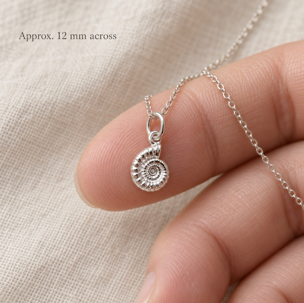 A silver spiral ammonite pendant necklace held on a person's fingers against a soft beige fabric background, with text reading 'Approx. 12 mm across'.