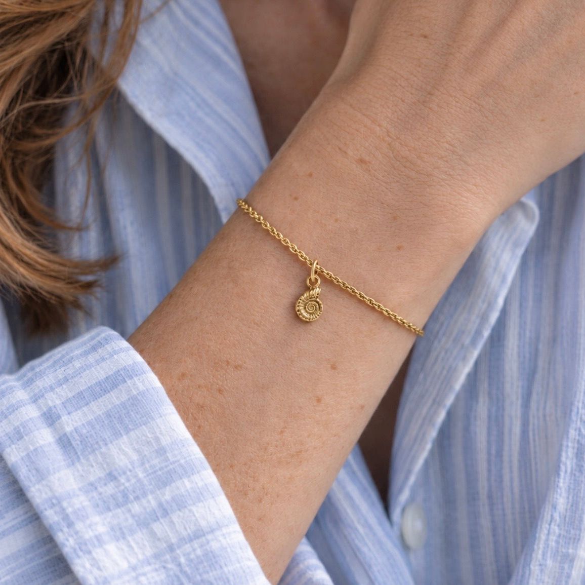 A delicate gold chain bracelet with a small ammonite fossil charm, shown worn on a person's wrist. The person is wearing a light blue and white striped button-down shirt.