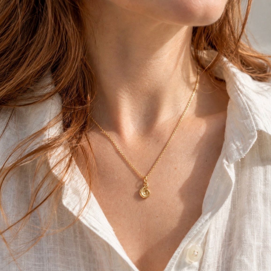 A close-up lifestyle shot of a woman wearing a delicate gold chain necklace with a small vermeil ammonite pendant, styled with a white linen shirt.