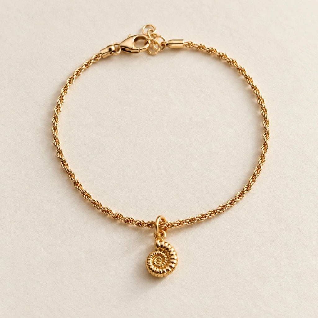 A delicate gold rope chain bracelet with a small ammonite charm, displayed on a light beige textured background.