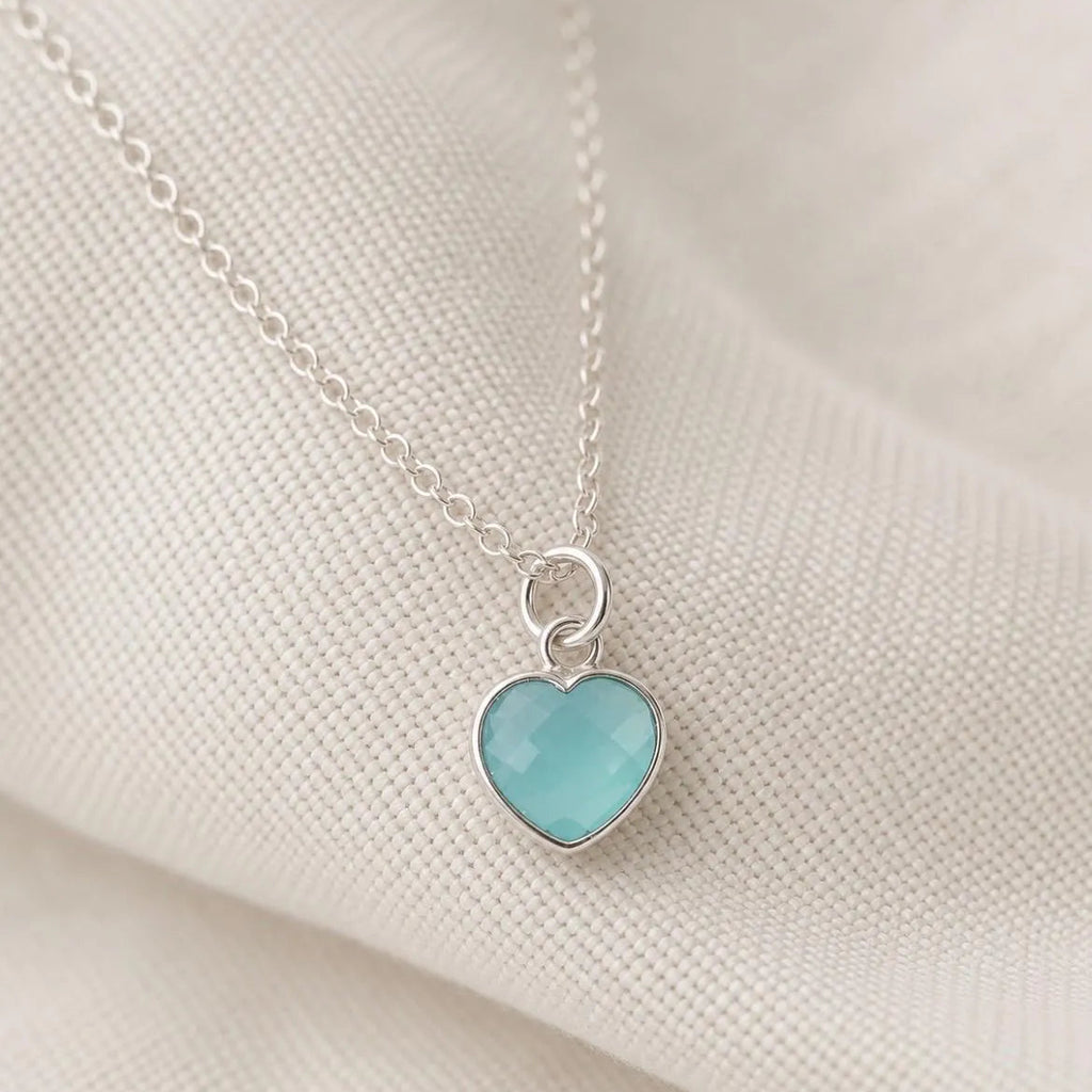 A silver heart pendant necklace with a light blue-green cabochon stone, resting on a textured cream fabric background.