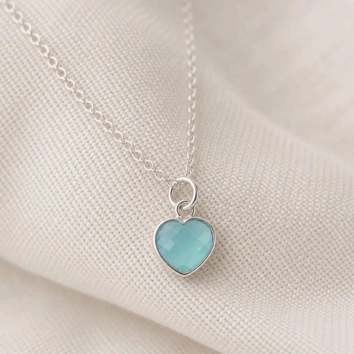A silver heart pendant necklace with a light blue-green cabochon stone, resting on a textured cream fabric background.