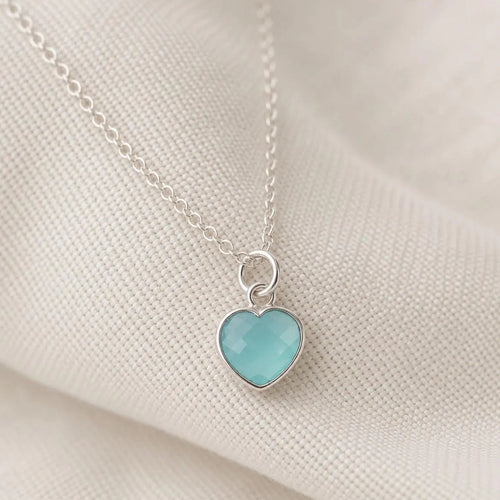 A silver heart pendant necklace with a light blue-green cabochon stone, resting on a textured cream fabric background.