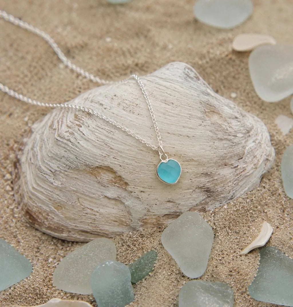 A silver chain necklace with a small heart-shaped turquoise pendant, displayed on a large weathered seashell over sand with scattered sea glass.