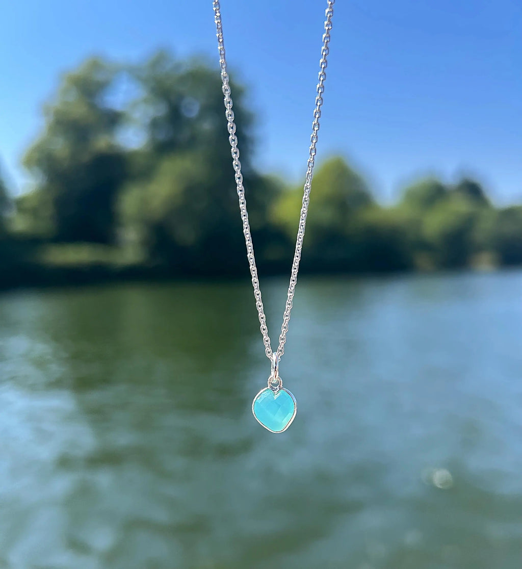 A silver chain necklace with a small heart-shaped turquoise pendant, photographed outdoors against a blurred background of water and trees.