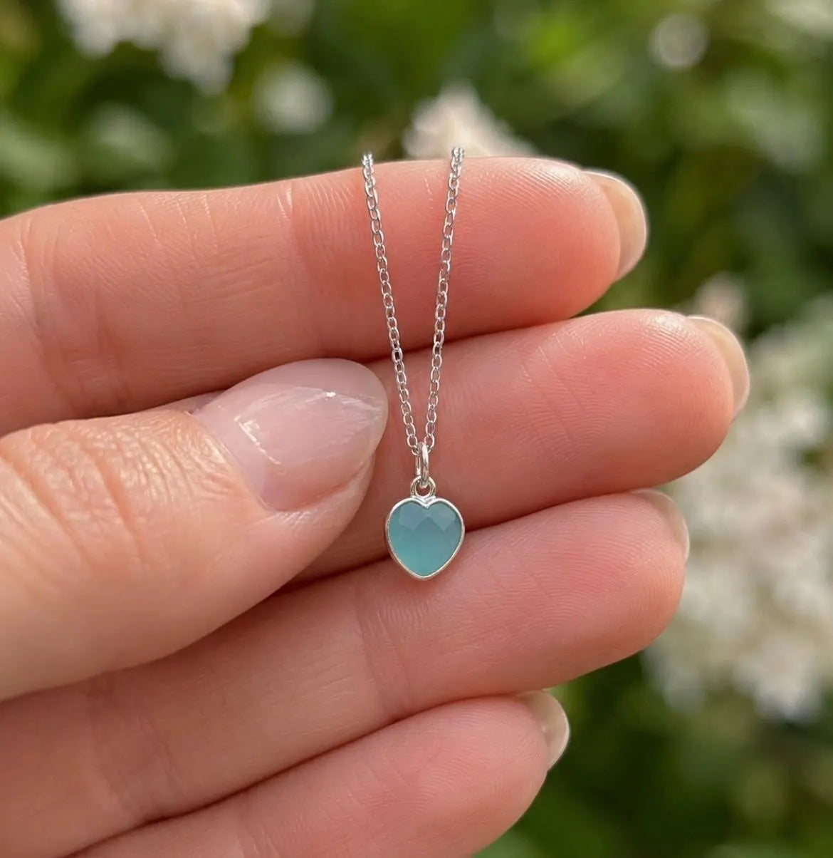 A silver heart pendant necklace with a light blue-green stone, held on a person's fingers against a blurred green outdoor background.