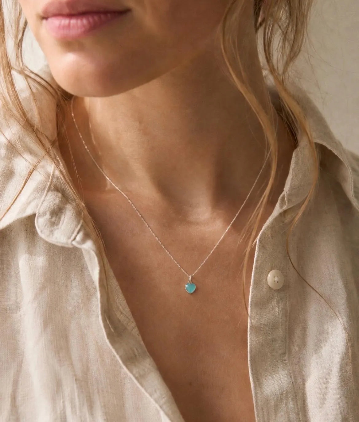 A close-up lifestyle shot of a woman wearing a delicate silver necklace with a small, round turquoise pendant, styled with a light beige linen shirt.