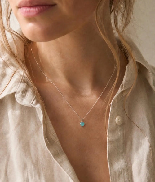 A close-up lifestyle shot of a woman wearing a delicate silver necklace with a small, round turquoise pendant, styled with a light beige linen shirt.