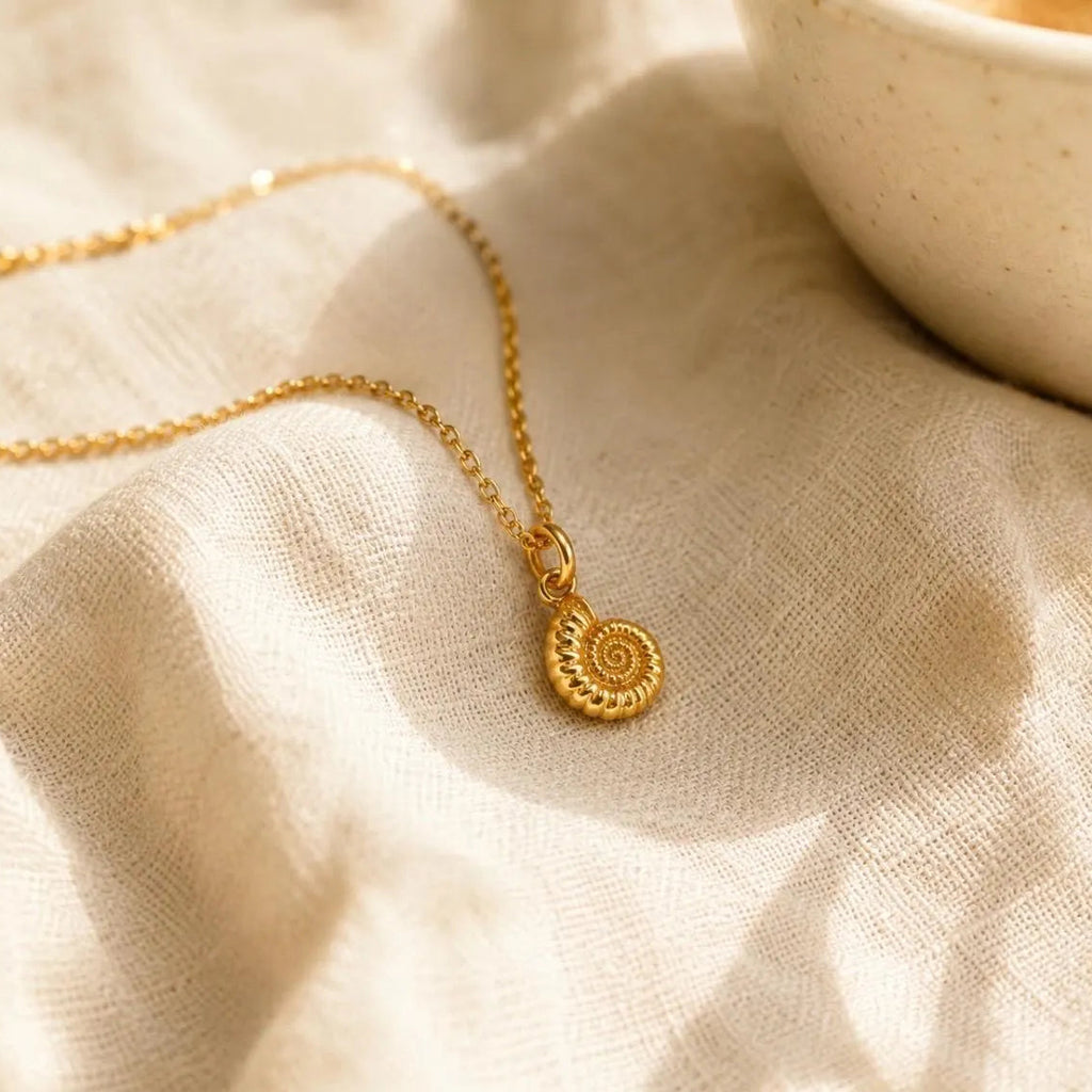 A delicate gold chain necklace with a small ammonite pendant, resting on a textured beige linen cloth with a ceramic bowl partially visible in the upper right.