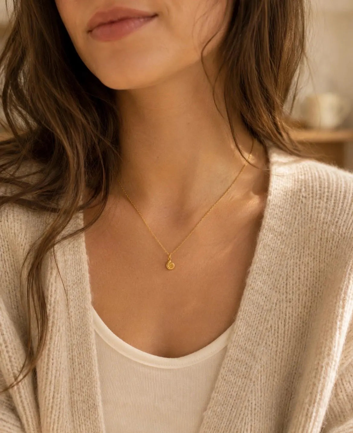 A close-up of a woman wearing a delicate gold necklace with a small teardrop-shaped pendant, styled with a cream ribbed tank top and a beige knit cardigan.
