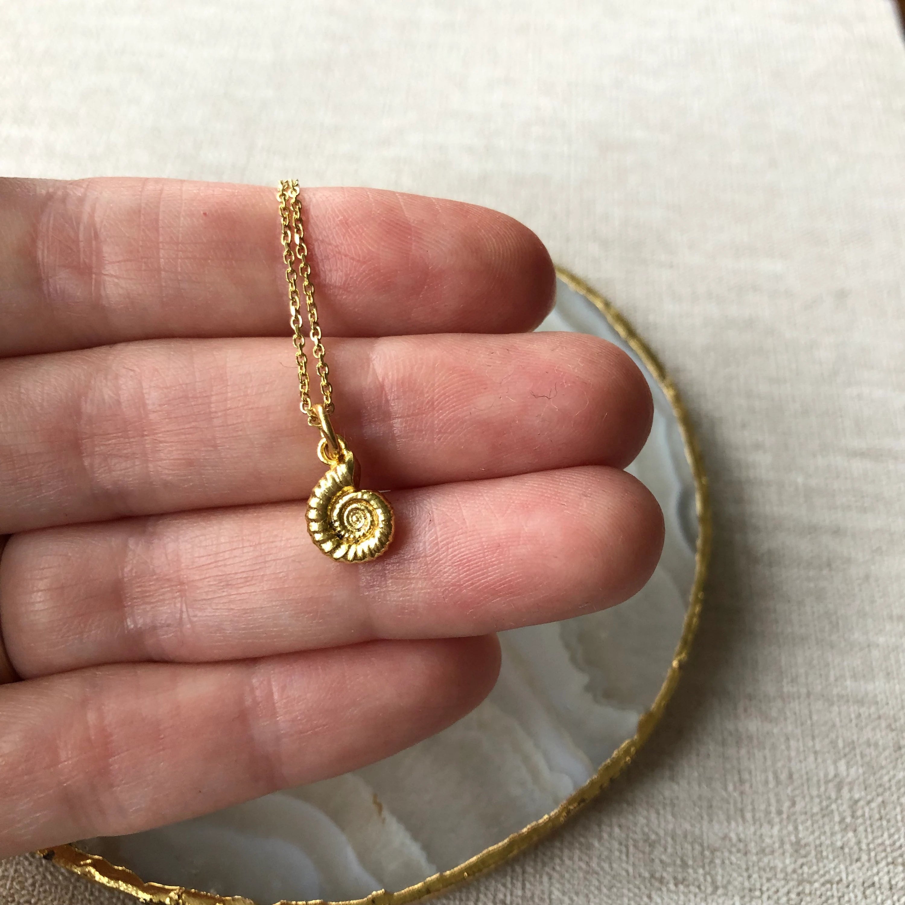 A gold necklace with a spiral shell pendant is held in a person's hand, with a textured gold bangle visible in the background on a light fabric surface.