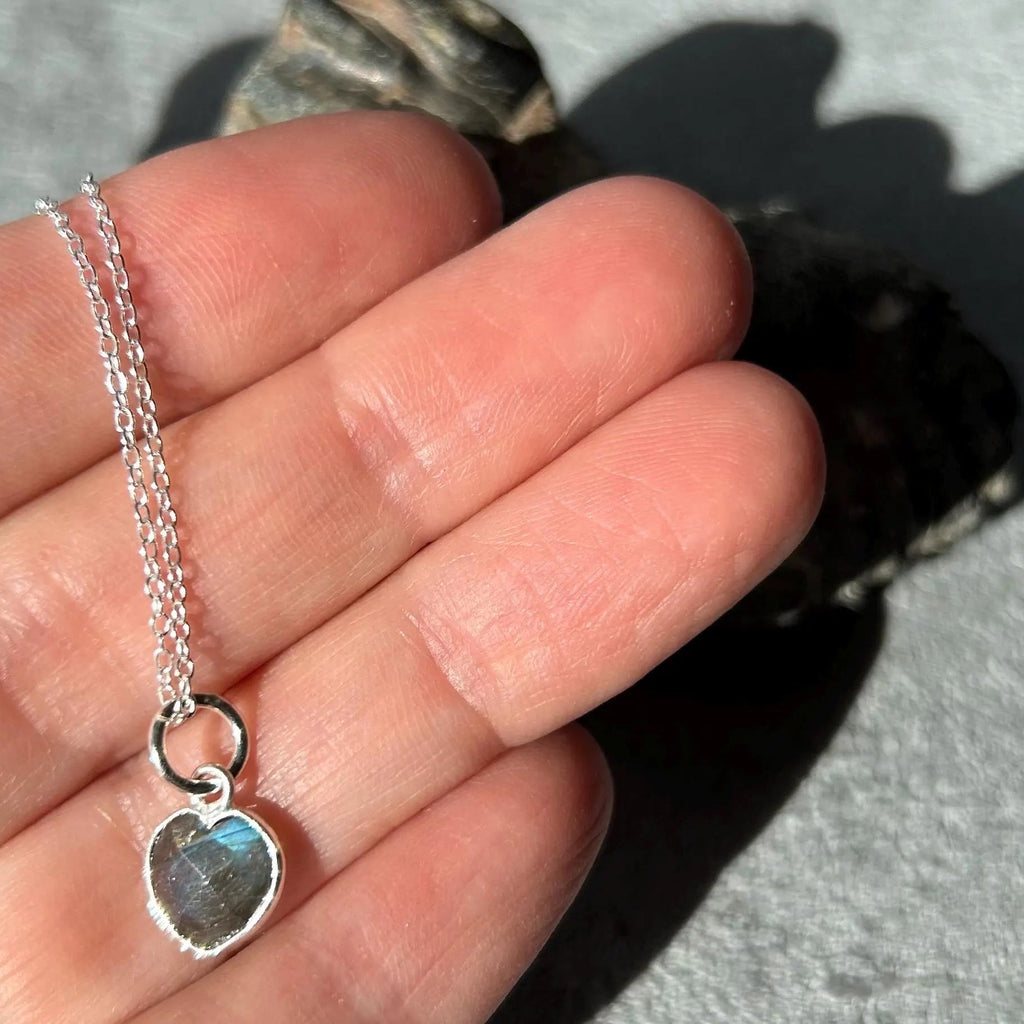 A sterling silver heart-shaped pendant necklace with a raw labradorite stone, shown resting on a person's fingers to demonstrate scale.