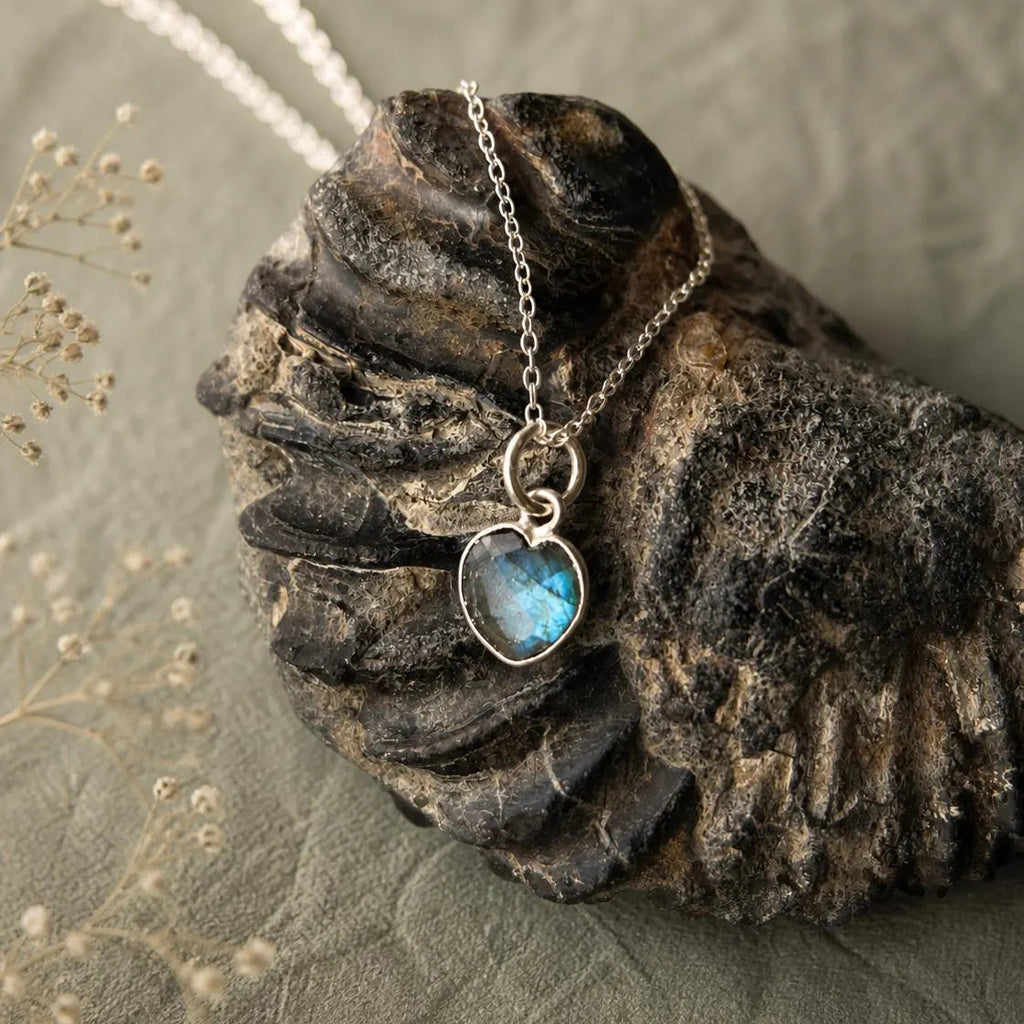 A silver heart-shaped pendant necklace with a blue-green iridescent stone, displayed on a dark textured shell over a neutral gray fabric background.