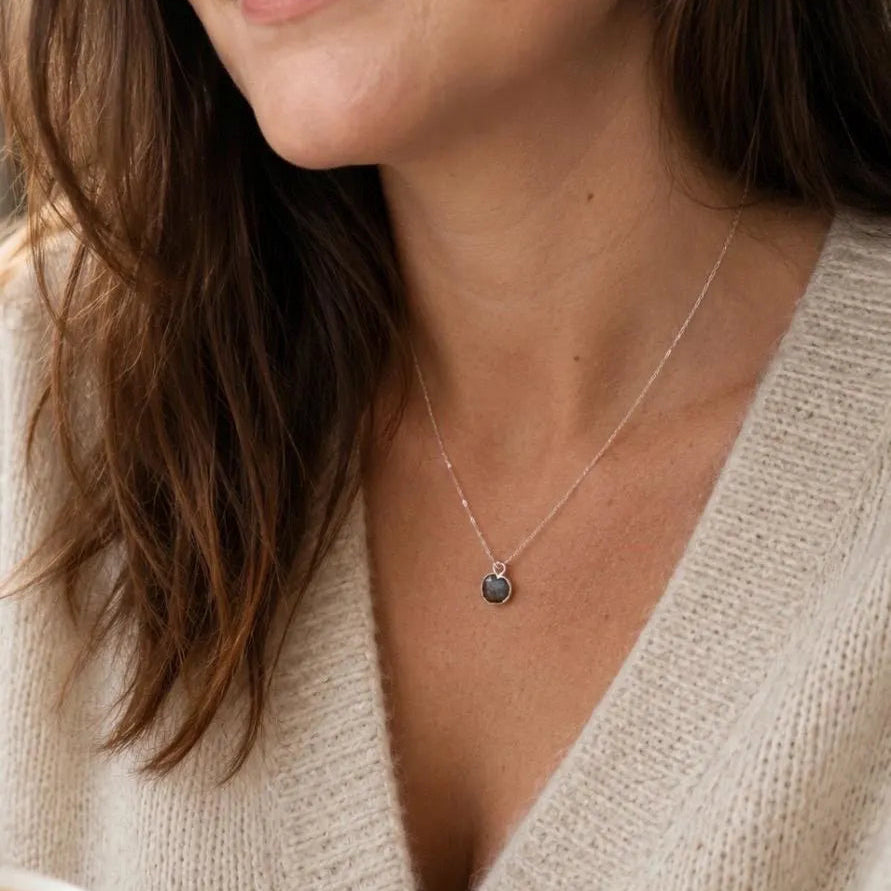 A close-up lifestyle shot of a woman wearing a delicate silver chain necklace with a small round dark gray stone pendant, paired with a cream-colored knit sweater.