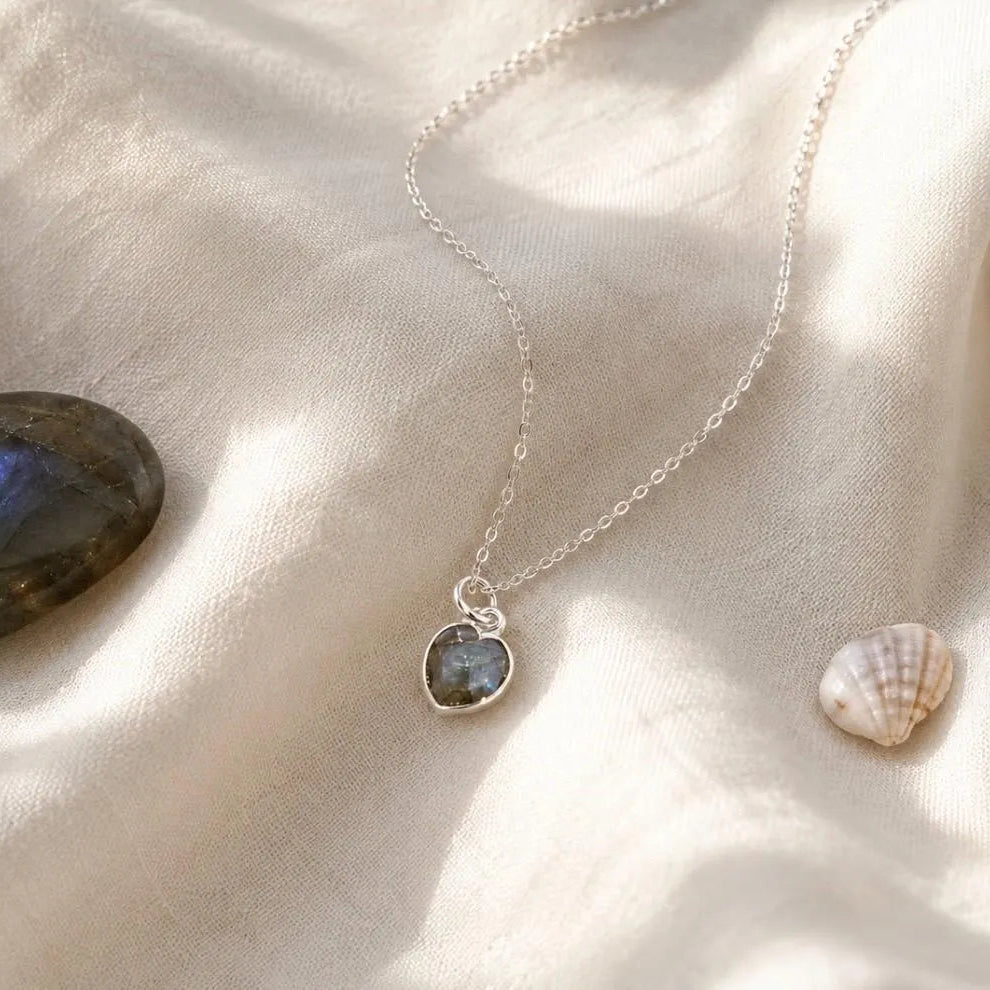 A silver chain necklace with a heart-shaped pendant featuring a dark grey to blue-grey labradorite stone, styled on draped cream satin fabric with a polished stone and a small seashell nearby.