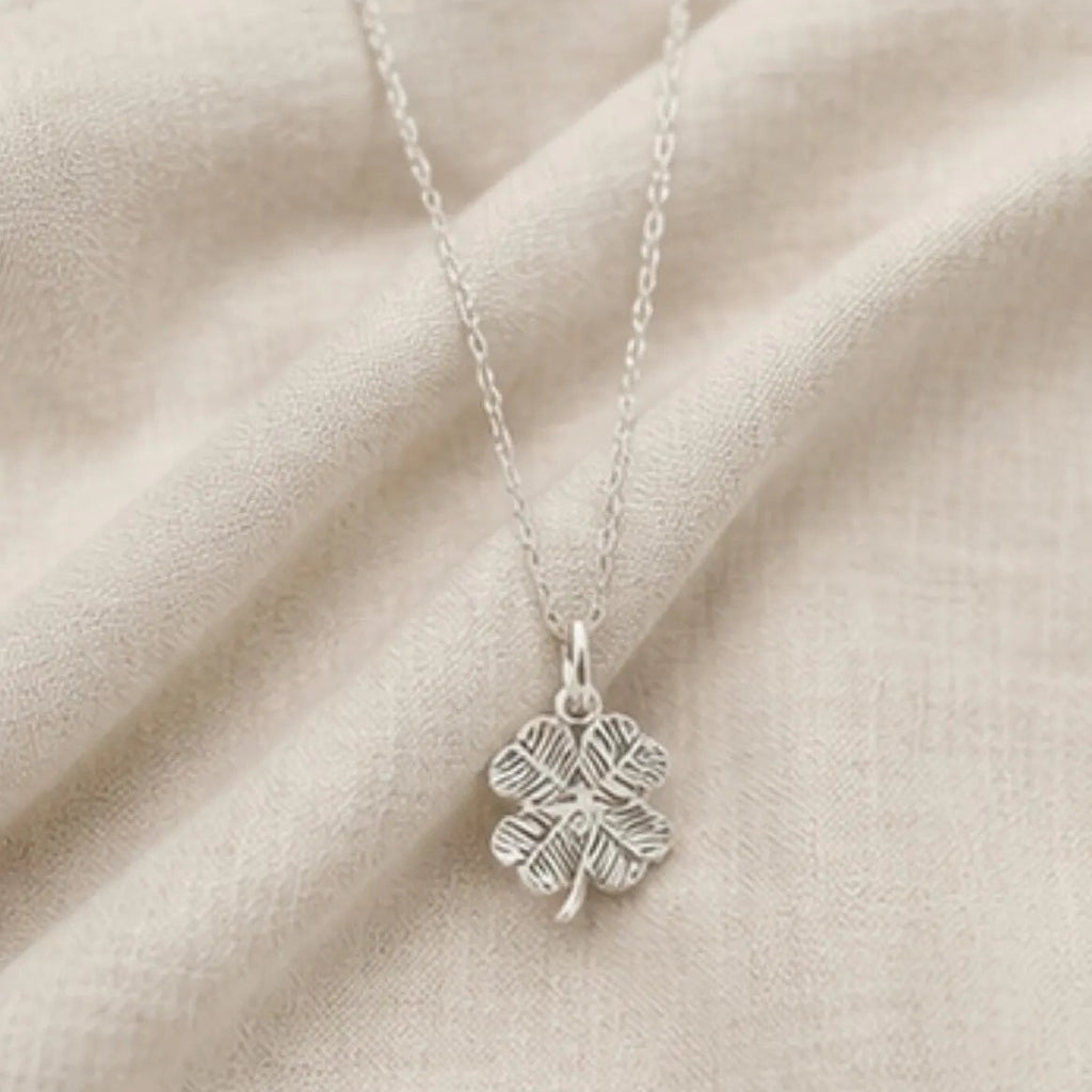 A sterling silver four-leaf clover pendant necklace with an oxidized textured finish, resting on a draped beige fabric background.