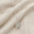 A sterling silver four-leaf clover pendant necklace with an oxidized textured finish, resting on a draped beige fabric background.