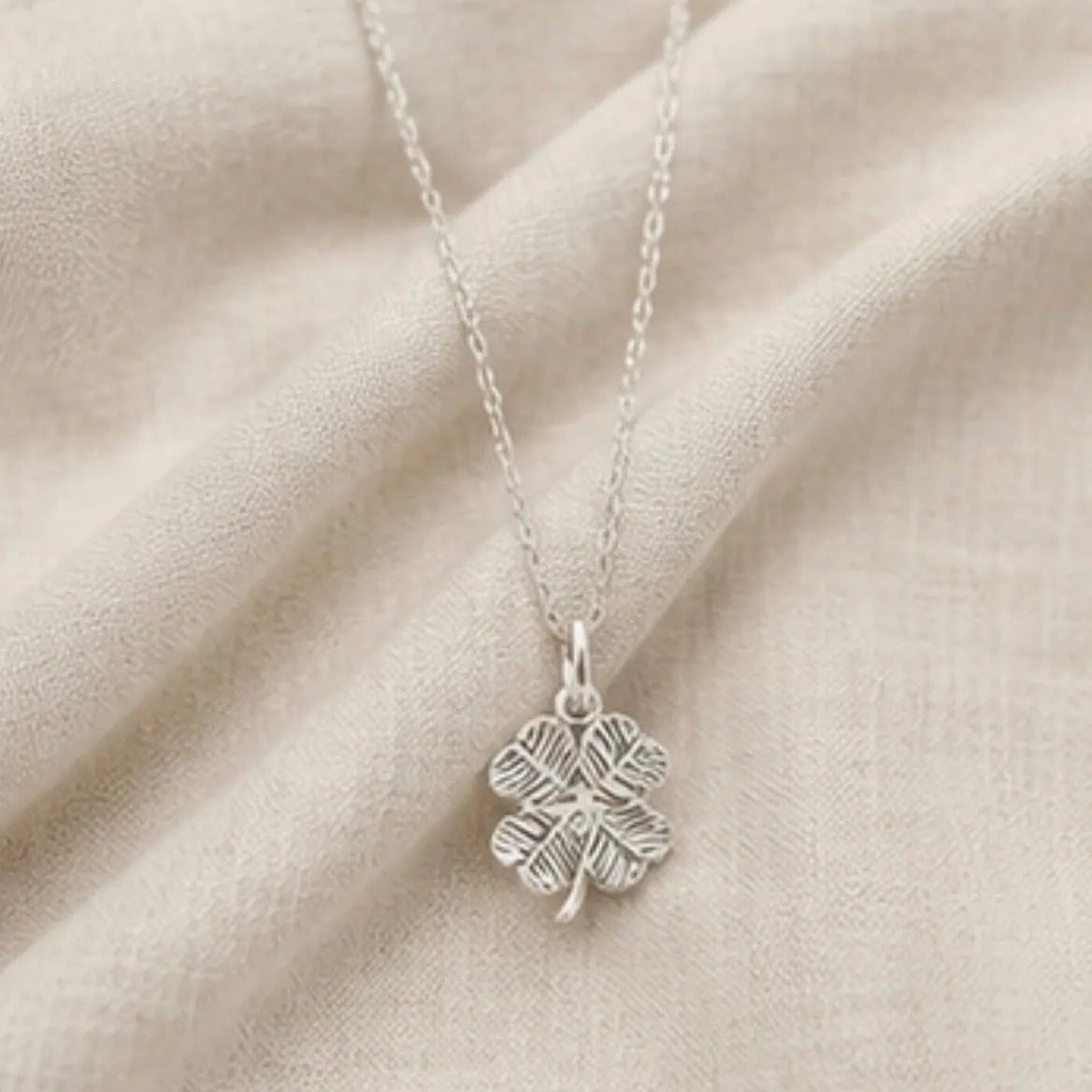 A sterling silver four-leaf clover pendant necklace with an oxidized textured finish, resting on a draped beige fabric background.