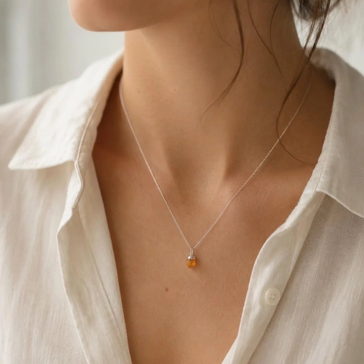 A close-up lifestyle shot of a woman wearing a delicate silver chain necklace with a small amber-orange bead pendant, paired with a white linen shirt.