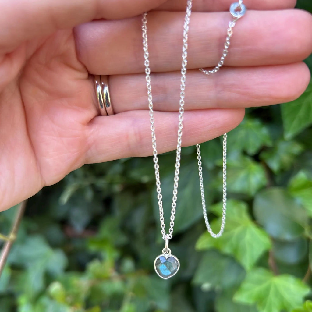 A silver chain necklace with a heart-shaped pendant containing a blue-green iridescent stone, held in a person's hand against a background of green ivy leaves.