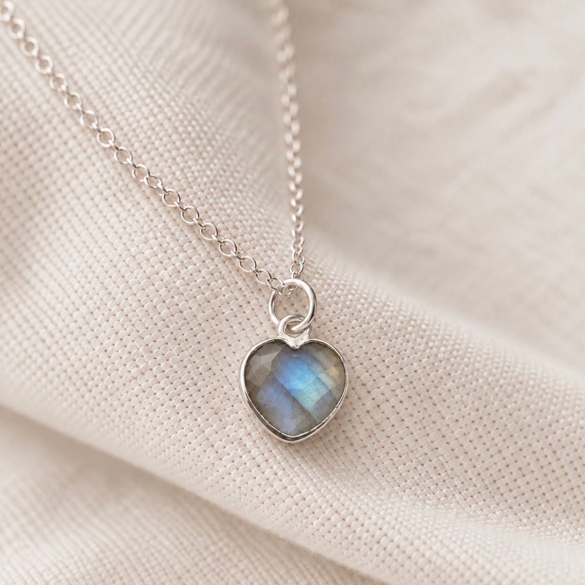A silver heart-shaped pendant necklace with a labradorite stone, resting on a textured cream-colored fabric background.