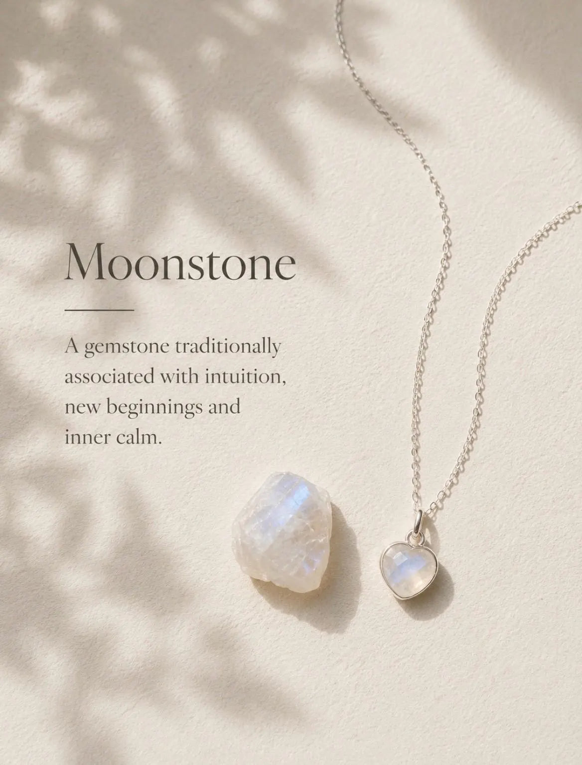 A silver necklace with a heart-shaped moonstone pendant is displayed on a warm cream background with soft leaf shadows. A larger raw moonstone gemstone is placed beside it, with the text 'Moonstone' and a descriptive quote on the left.