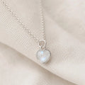 A silver heart pendant necklace with a milky white iridescent stone, resting on a textured beige fabric background.