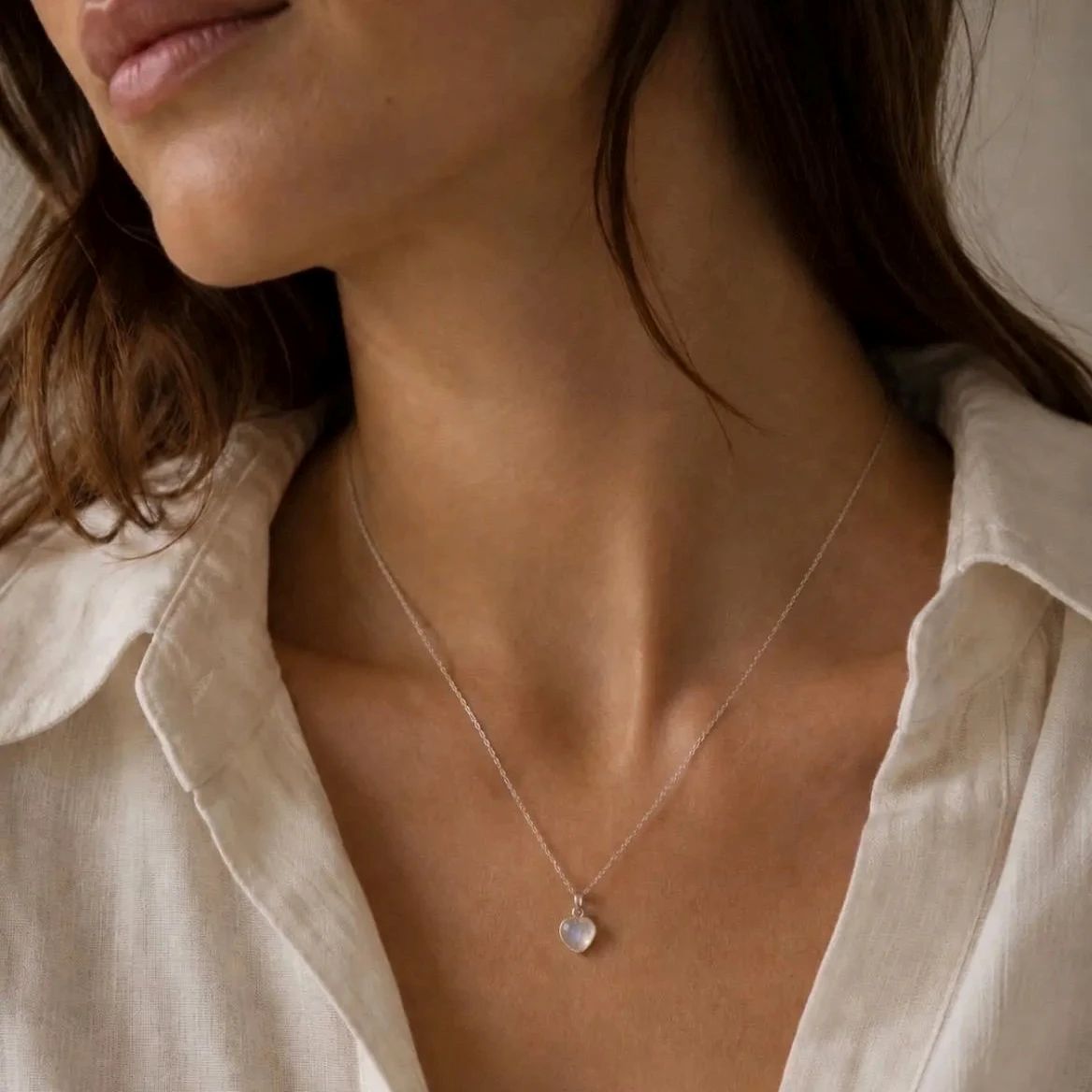 A close-up of a woman wearing a delicate silver chain necklace with a small, heart-shaped clear moonstone stone pendant, paired with a light beige linen shirt.