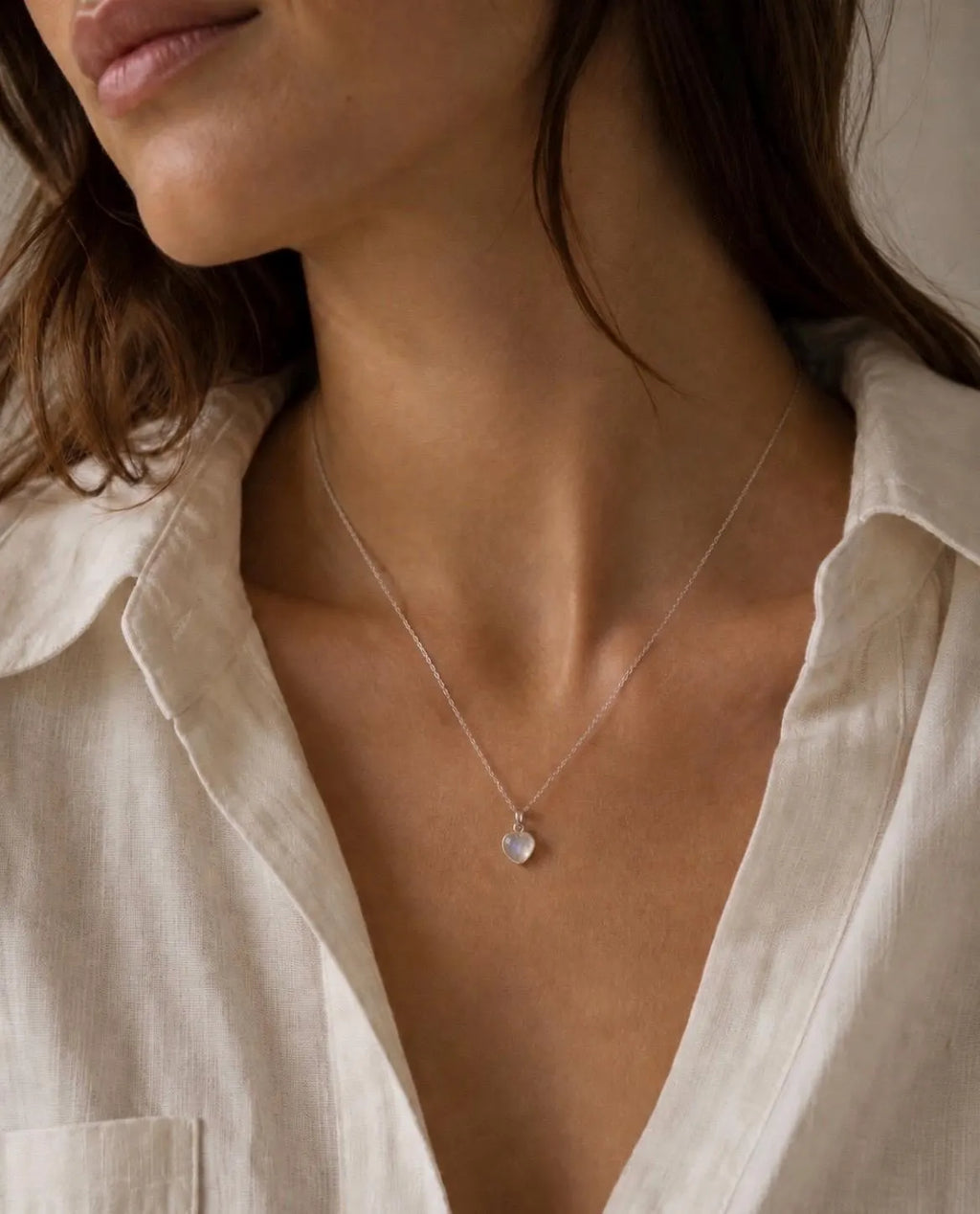 A close-up of a woman wearing a delicate silver chain necklace with a small, clear teardrop-shaped crystal pendant, styled with a light beige linen shirt.