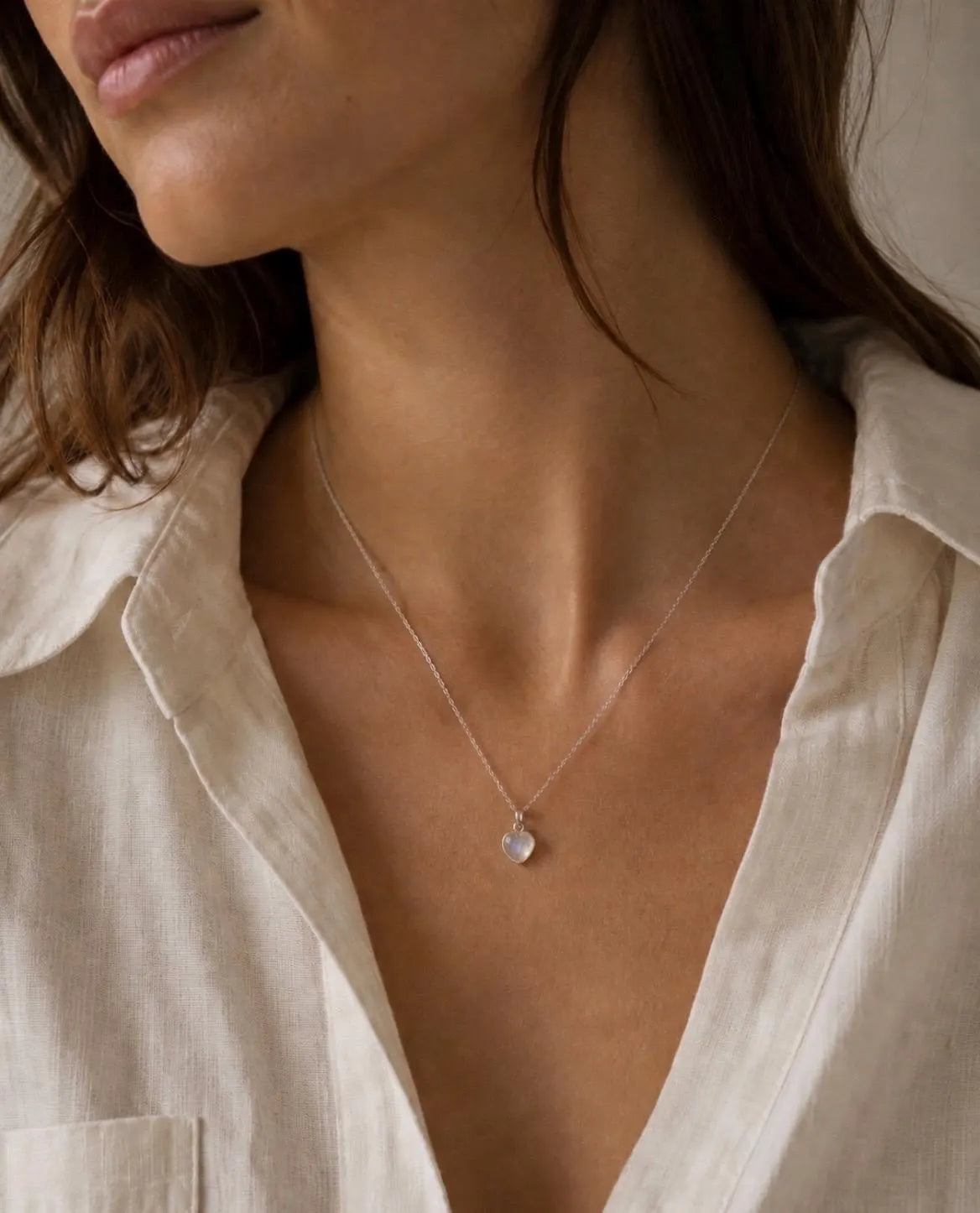 A close-up of a woman wearing a delicate silver chain necklace with a small, clear teardrop-shaped crystal pendant, styled with a light beige linen shirt.