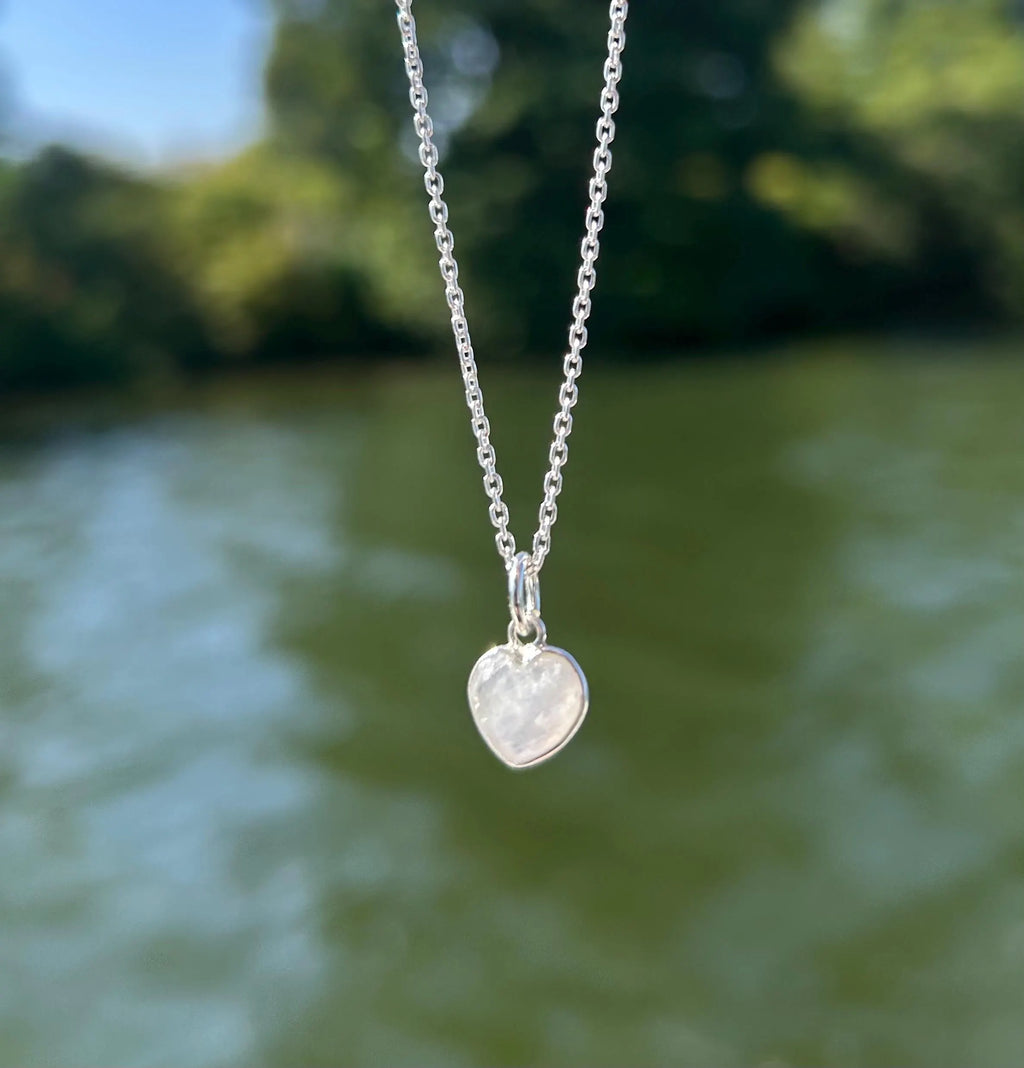 A silver heart pendant necklace with a milky white iridescent stone, hanging against a blurred outdoor background of green foliage and blue water.