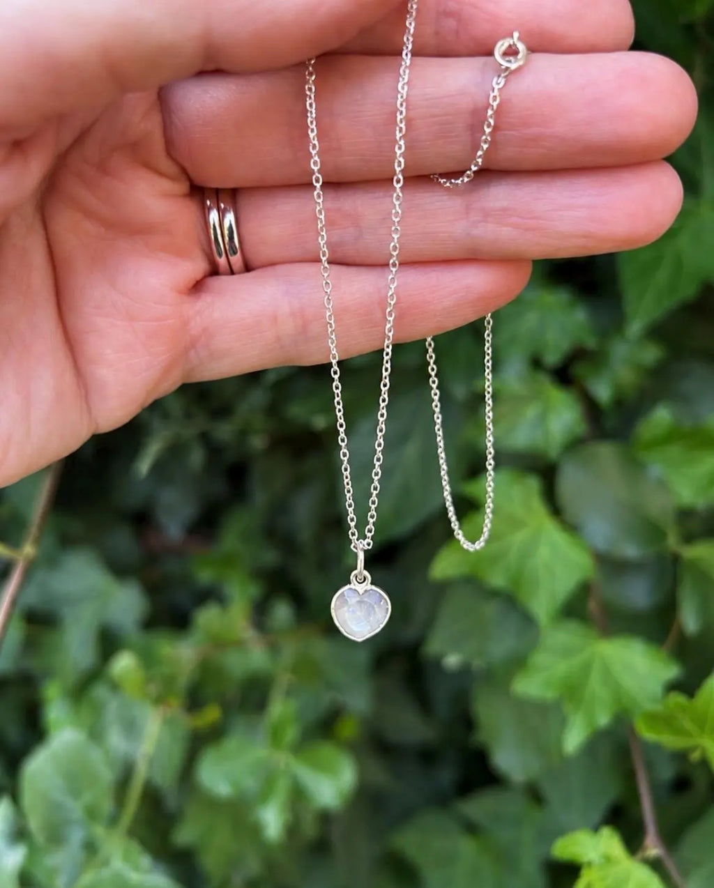 A delicate silver chain necklace with a small heart-shaped pendant is held in a person's hand, with green ivy leaves blurred in the background.