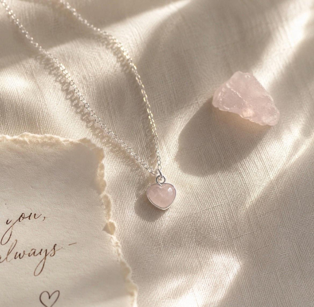 A delicate silver chain necklace with a small heart-shaped pink stone pendant, styled on draped cream fabric alongside a torn paper note and a rough pink stone.