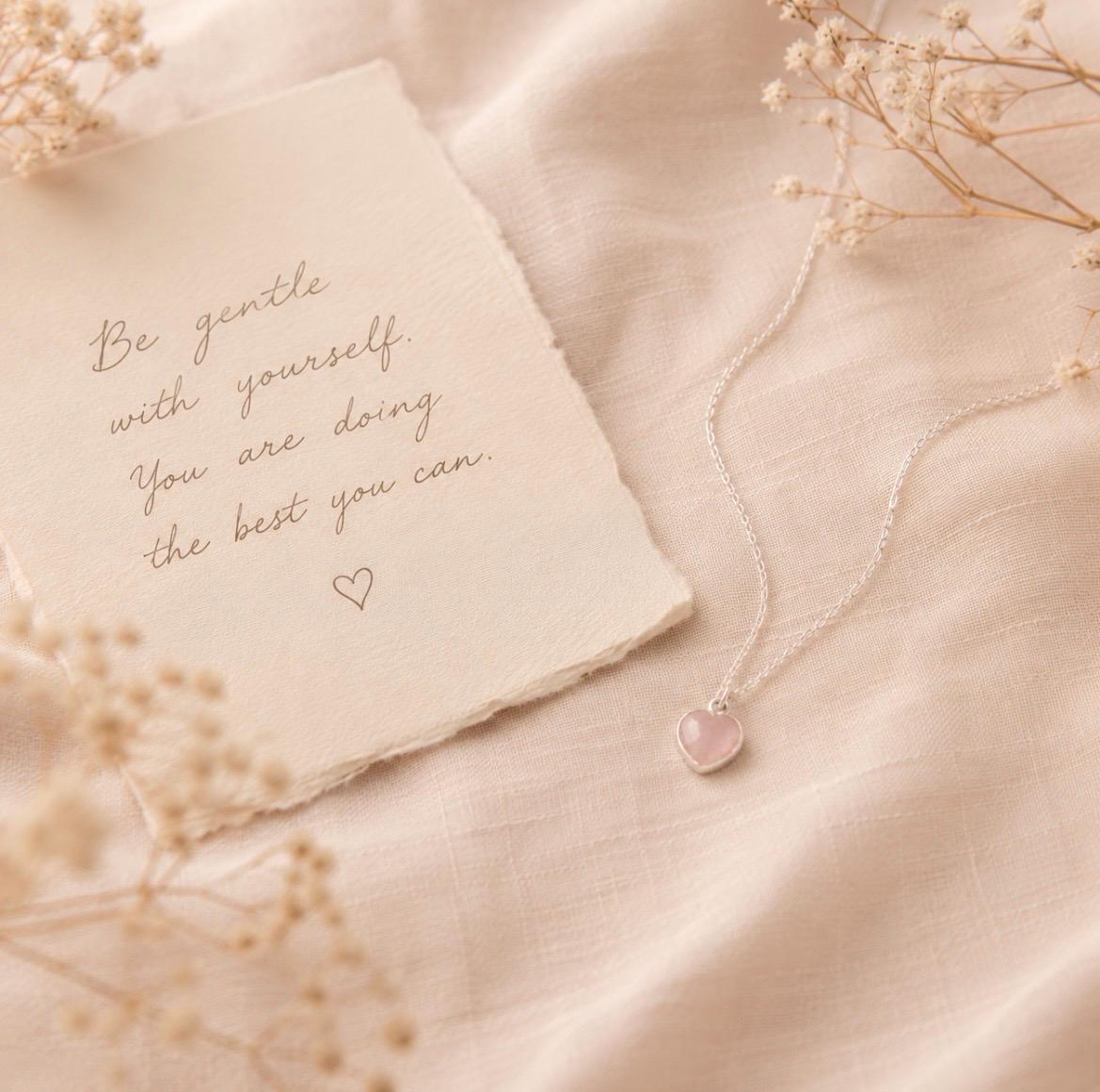 A delicate silver necklace with a small pink heart pendant is arranged on soft cream fabric beside a torn paper note with cursive text and dried flowers.