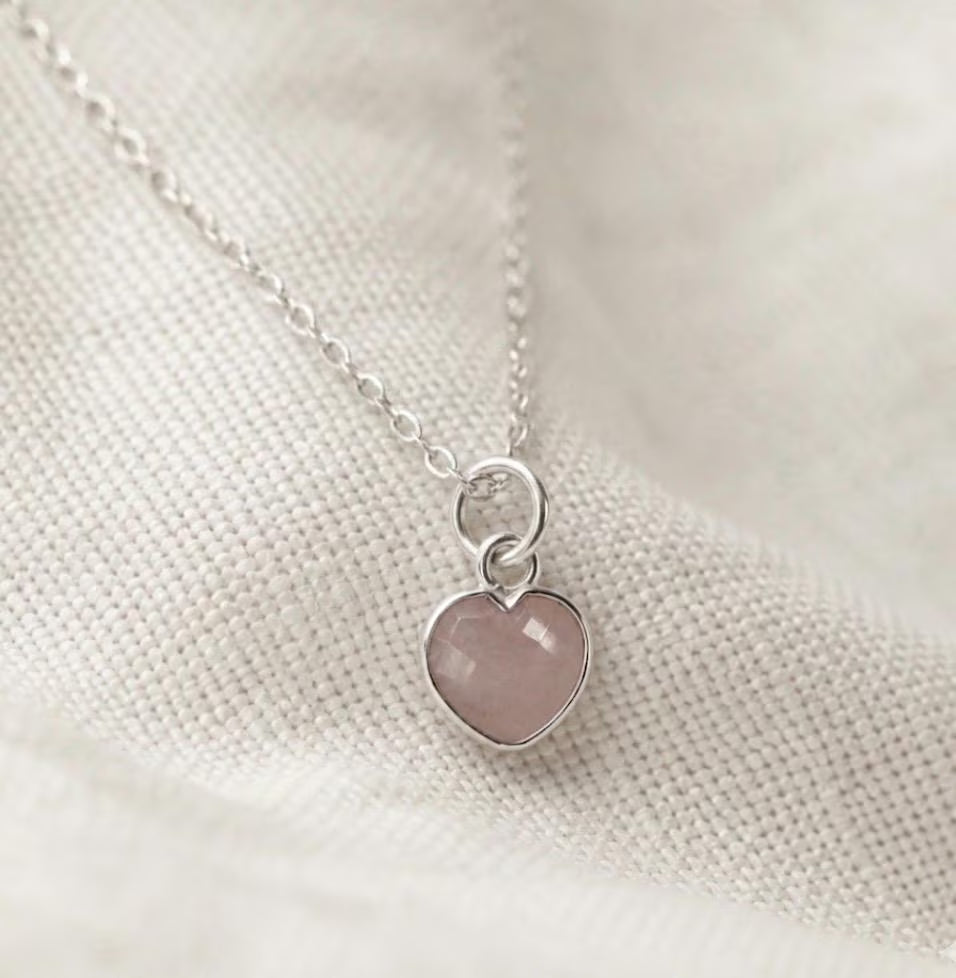 A delicate silver chain necklace with a small heart-shaped pendant featuring a smooth, rose quartz stone, resting on a textured cream fabric background.