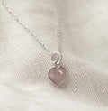 A delicate silver chain necklace with a small heart-shaped pendant featuring a smooth, rose quartz stone, resting on a textured cream fabric background.