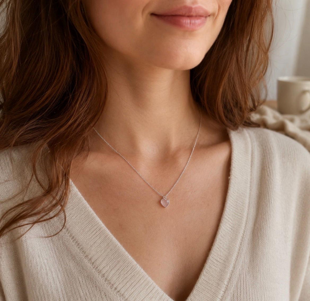 A close-up lifestyle shot of a woman wearing a delicate silver chain necklace with a small, pale pink heart-shaped pendant. She is wearing a cream-colored V-neck sweater, and a soft, neutral-toned mug is visible in the background.