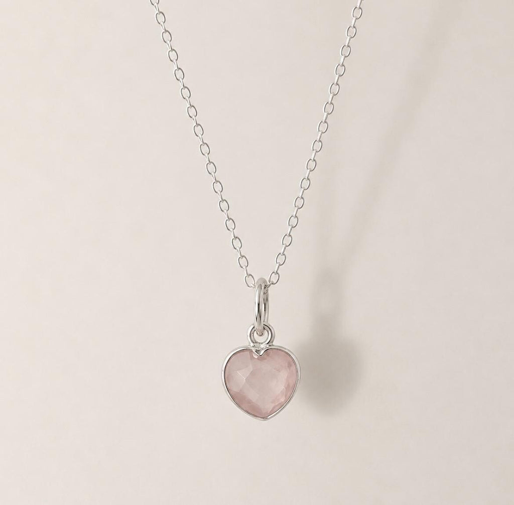 A delicate silver chain necklace with a small heart-shaped pink rose quartz gemstone pendant, displayed against a soft, light beige background.