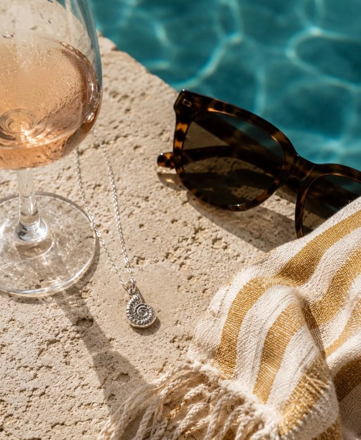 A silver pendant necklace with a spiral design is displayed on a textured beige stone surface next to a glass of rosé wine, tortoiseshell sunglasses, and a cream and gold striped towel, with a swimming pool in the background.