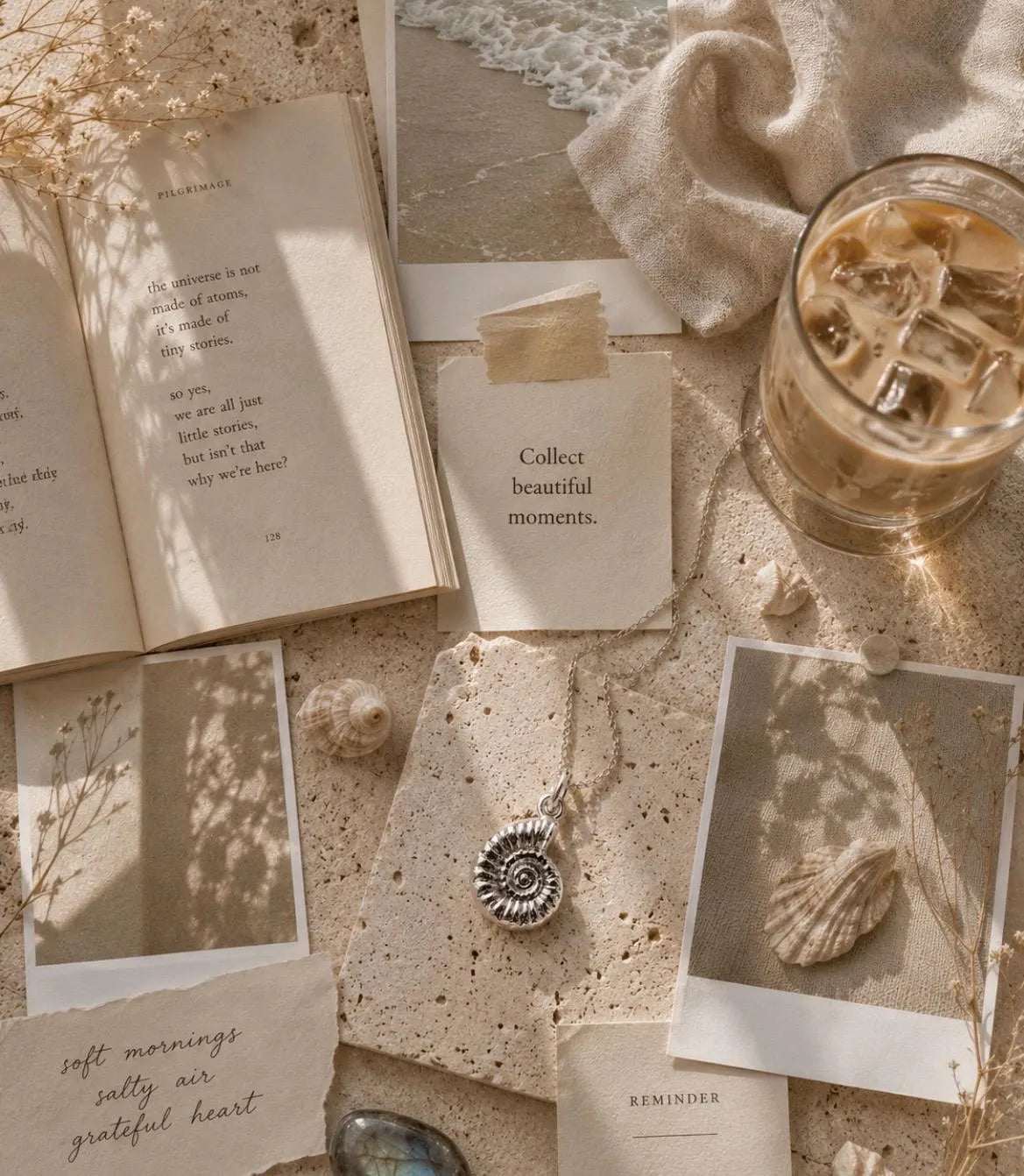 A styled flat-lay featuring a silver spiral ammonite pendant necklace on a textured beige surface, surrounded by an open book, a glass of iced coffee, seashells, and cards with boho-style text.