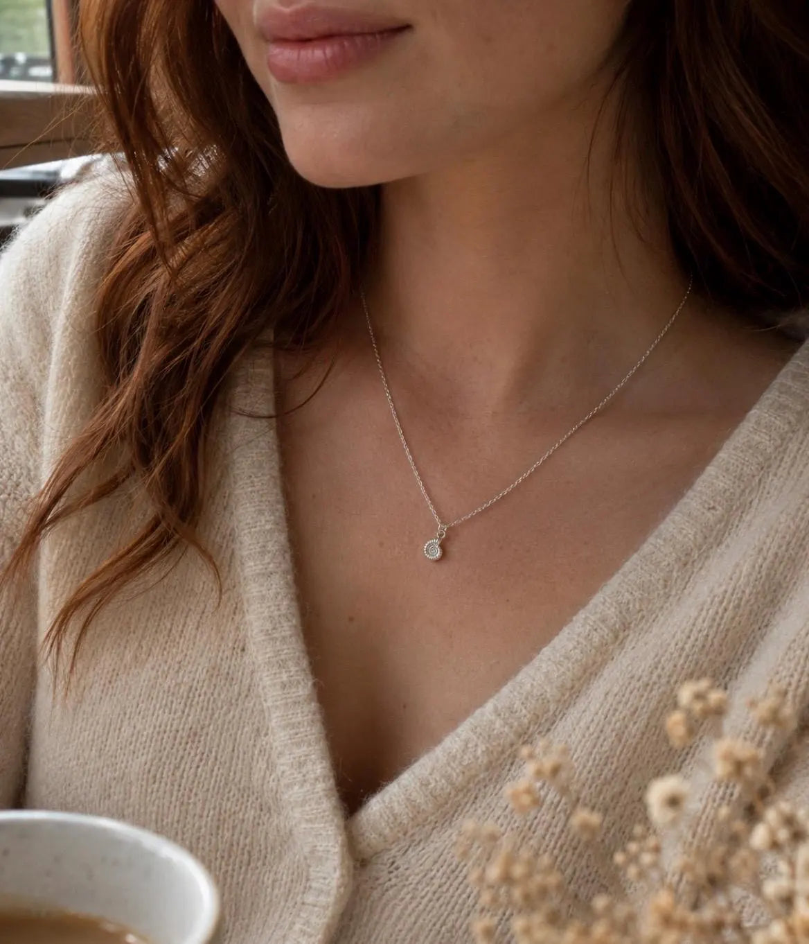 A close-up lifestyle shot of a woman wearing a delicate silver chain necklace with a small ammonite pendant, styled with a beige knit cardigan.