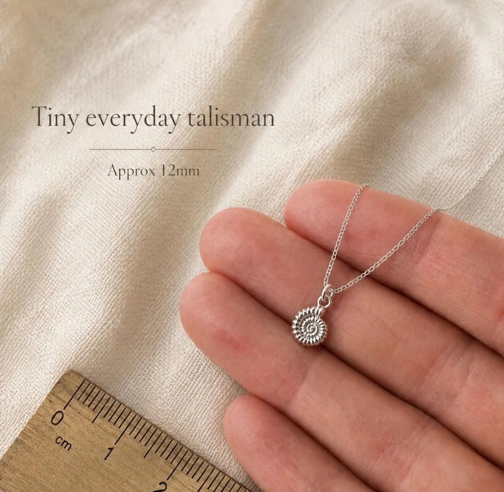 A silver necklace with a small spiral ammonite pendant is held on a person's fingers over a cream fabric background, with text reading 'Tiny everyday talisman' and 'Approx 12mm' plus a wooden ruler for scale.